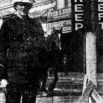 Image: A Caucasian man in an early twentieth century police uniform stands next to a marker with the words ‘Keep’ and ‘Left’ painted on it in large block letters