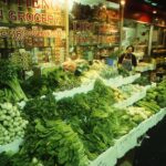 Image: woman standing with large display of vegetables in front of Asian grocery store