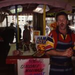 Image: man standing in front of market stall, caught mid shout