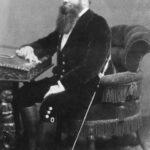 Image: a bearded man in three quarter profile and wearing a hat and sword sits at his writing desk.