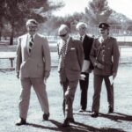 Image: Four men walk together as a group along the periphery of a parkland. Three are dressed in suits while the fourth wears a military-style uniform