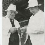 Image: Two men in white coats and hats smiling and engaging in conversation