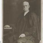 Image: Man dressed in robes, with large white moustache and glasses sits on a chair by a desk with his hand on a bible