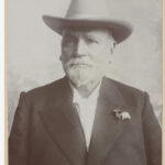 Image: A photographic portrait of an elderly Caucasian man with a white goatee. He is wearing a Stetson hat and a dark-coloured overcoat