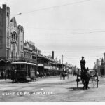 Busy street with several horse drawn carts and one electric tram car, a large building dominates the streetscape