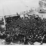 Image: a large crowd of workers gather for a strike at a wharf, the ocean can be seen in the background