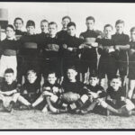 Image: a group of boys in football uniforms with their teacher. The boy seated in the centre of the front row holds a football with