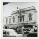 Image: Front view of a theatre building with cars driving in the foreground and people on the street