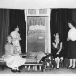 Image: Two women sitting, and two women standing on a stage with a dark curtain backdrop and various stage props