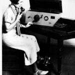 Image: A Caucasian woman in a 1930s-era dress sits at a table upon which sits a large radio set. The woman wears a headset and holds the radio receiver