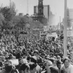 Image: a group of people in 1970s clothing gather in a city square holding protest banners against the Vietnam War.