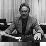 Image: Black and white photograph of a smiling man in jacket sitting at his desk holding a sheet of paper