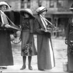 Image: Three women and a young girl wearing hats and long coats stand in the street holding sprigs of wattle plant