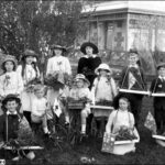 Image: A group of children stand near a house holding trays of flowers, two are on bicycles, and some are holding Australian flags