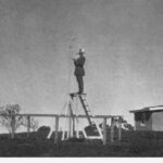 Image: A man, smoking a pipe, stands on a ladder collecting data at an outside, rural weather observatory