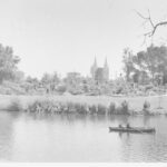 Image: Two men sitting in a row boat on a River, with a park and Cathedral visible in the background