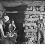 Image: Three men working inside a mine shaft with stacked timber walls and ceiling, and two trolleys filled with stone materials